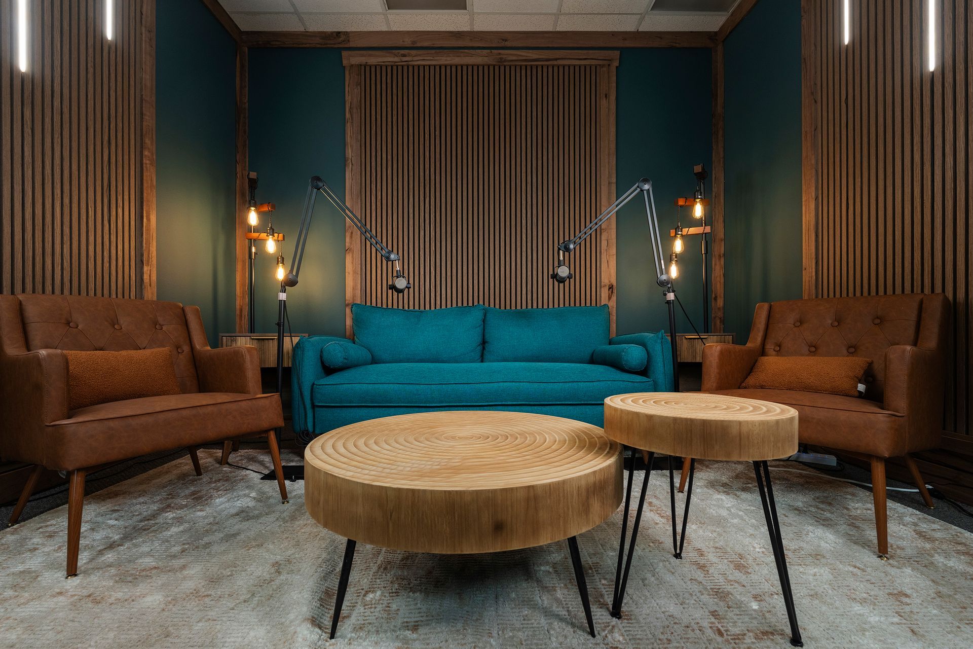Modern lounge with teal sofa, brown chairs, round coffee tables, and wall sconces against wood paneling.