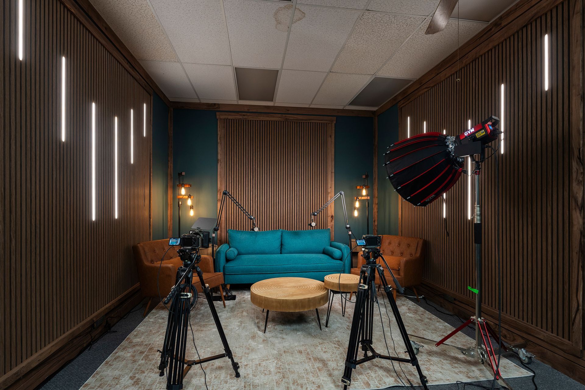 Studio set with teal couch, coffee table, tripods, lights, and curtains in a wood-paneled room