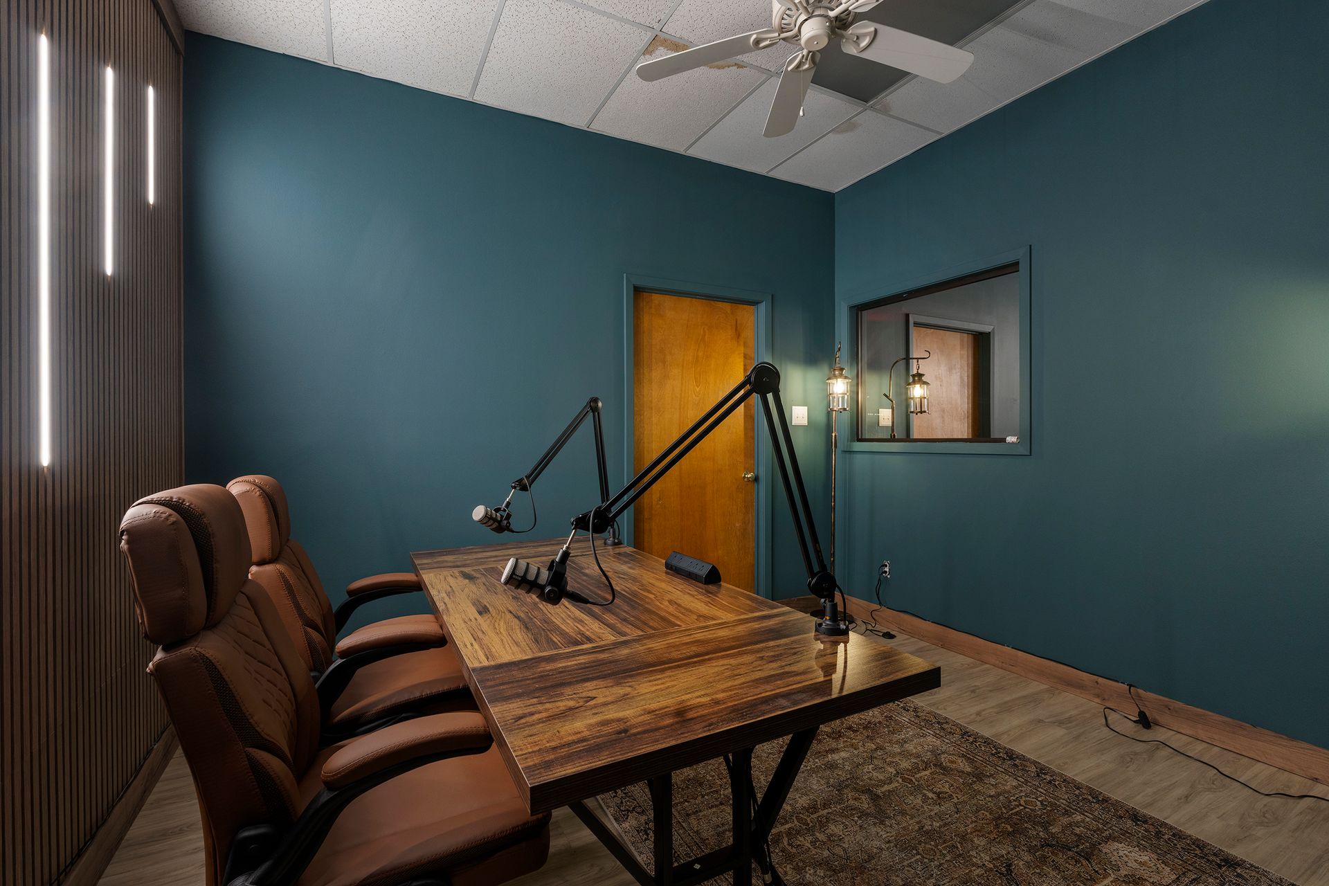 Podcast studio with teal walls, wooden table, microphones, brown chairs, and a window into a recording booth