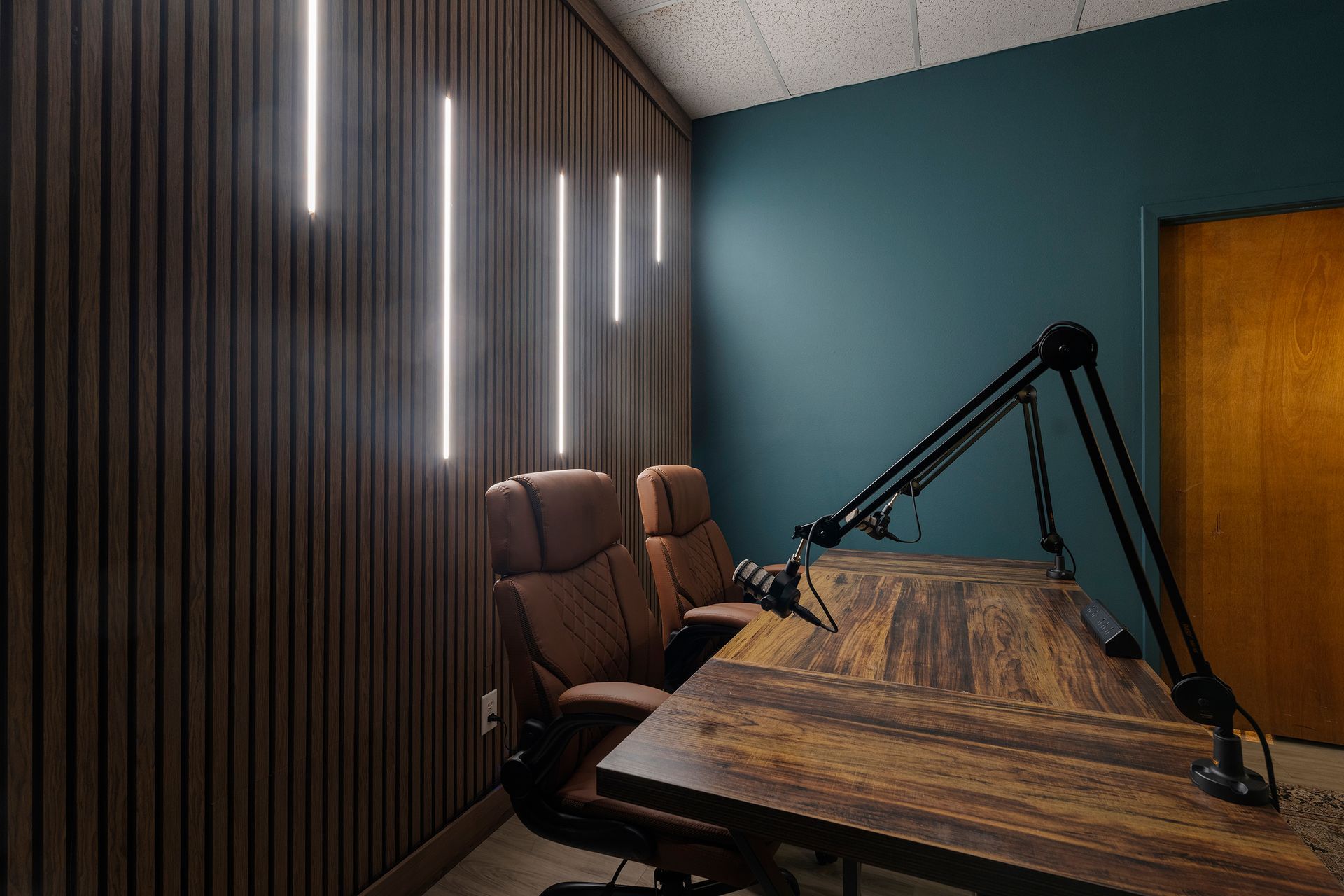 Modern podcast studio with wooden table, brown chairs, and black microphone arms against a wood and teal wall