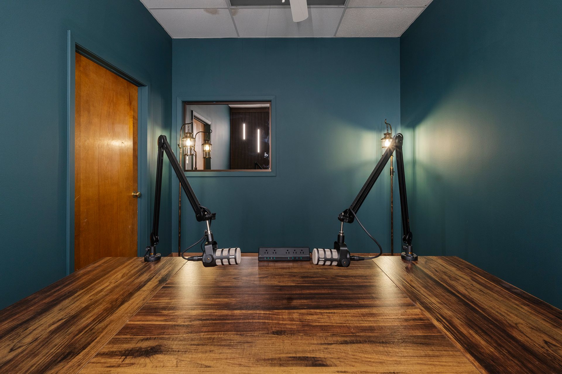 Empty recording studio with teal walls, wood floor, microphones on stands, and a small window.