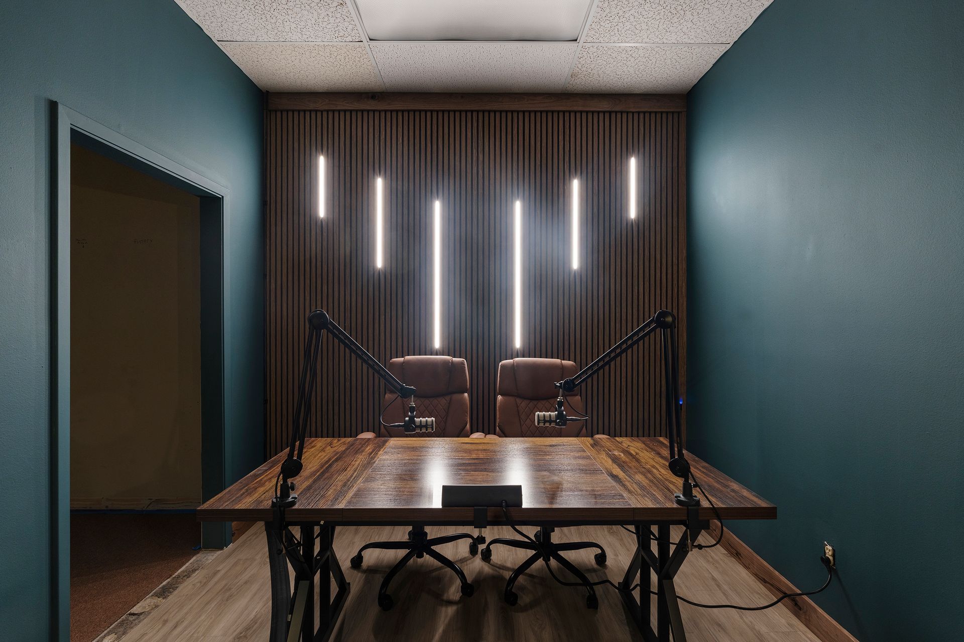 Small podcast studio with two microphones, wooden table, and vertical LED lights on a dark accent wall