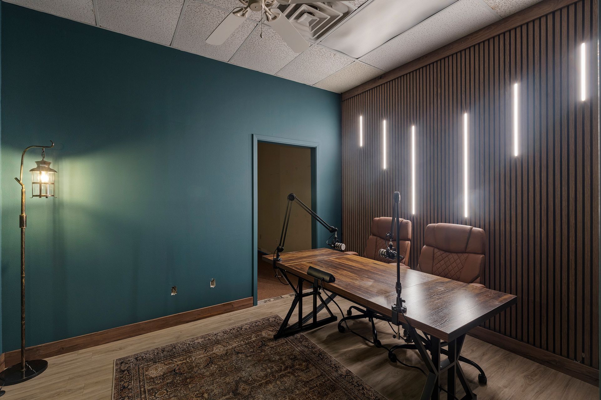 Minimal recording studio with teal wall, vertical lights, couch, and a table with microphone stands.