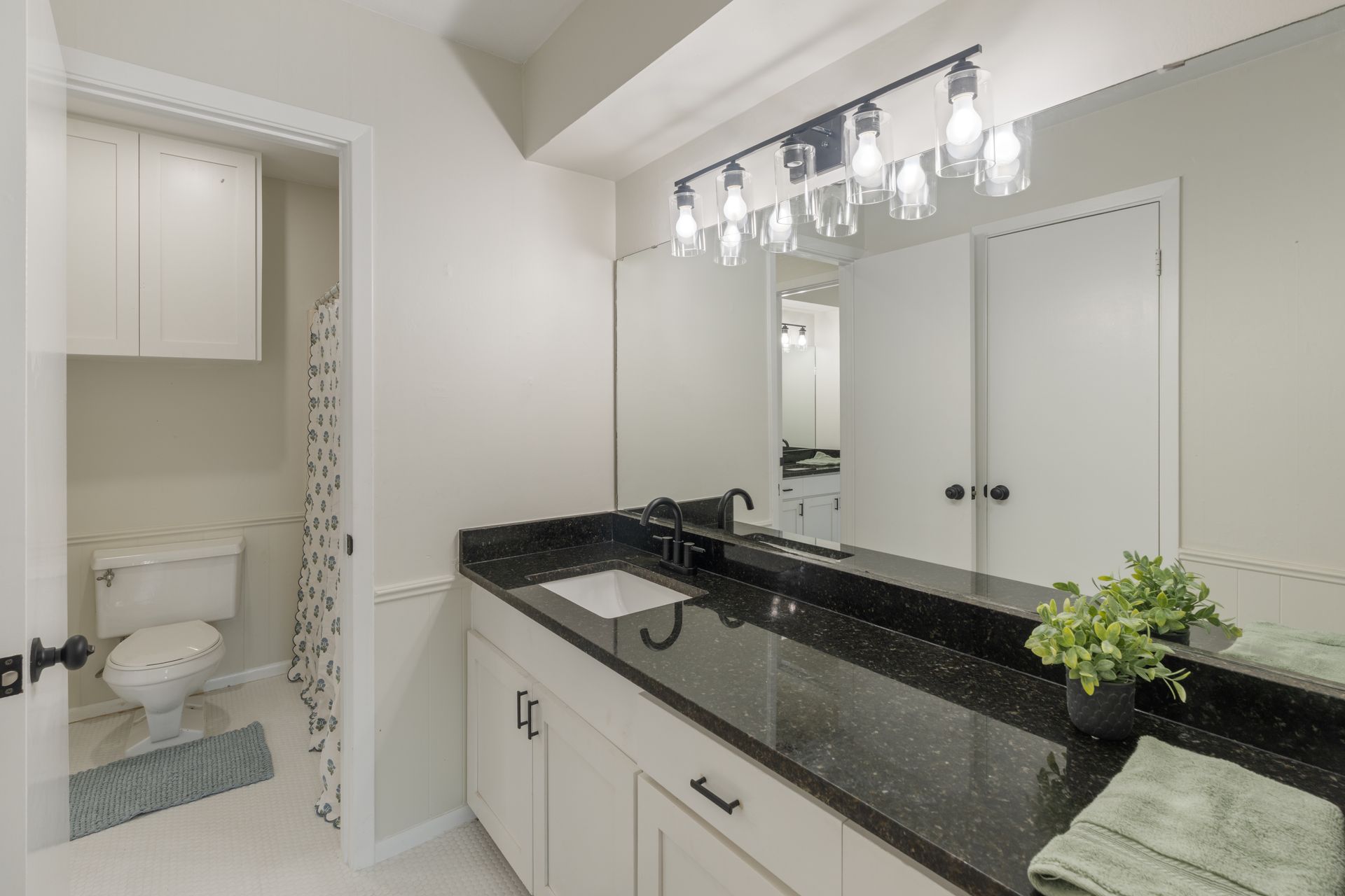 Modern bathroom vanity with black countertop, large mirror, and separate toilet room visible through open door