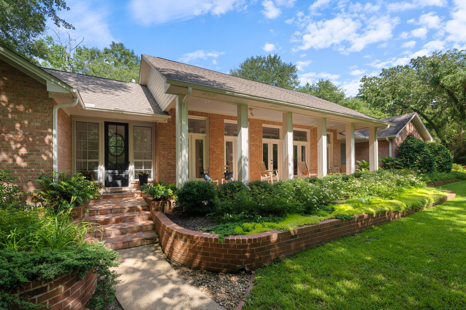 Brick house with white columns, front porch, and lush green landscaping under a blue sky