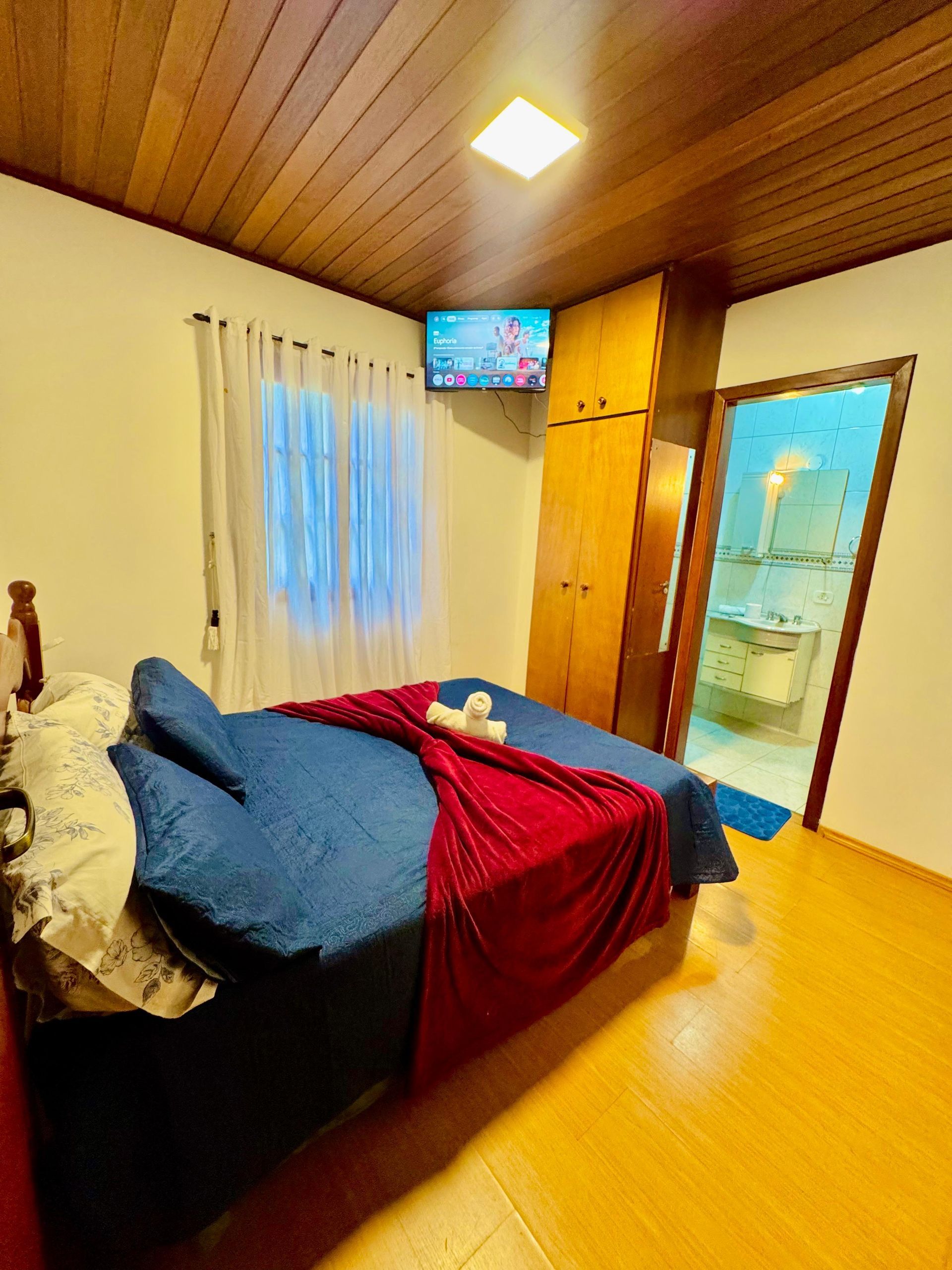 Quarto aconchegante com colcha azul, cobertor vermelho, TV de parede, teto de madeira e porta aberta para o banheiro.