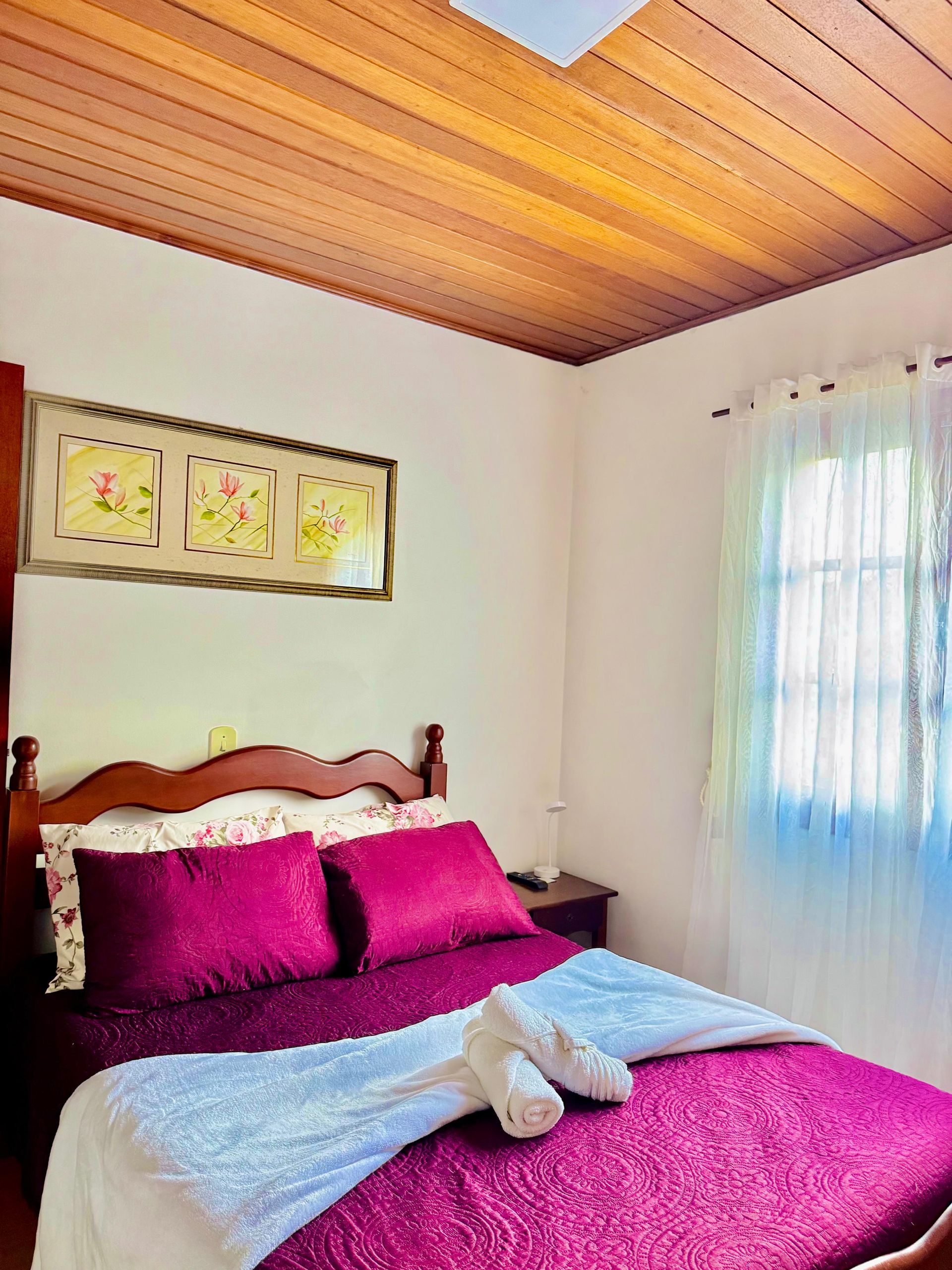 Quarto aconchegante com teto de madeira, colcha rosa, quadros e cortinas azul-claras junto à janela.