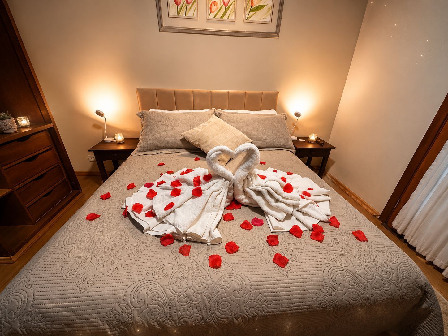 Cama romântica com cisnes de toalha branca, pétalas de rosa vermelha e abajures com luz quente em um quarto aconchegante.