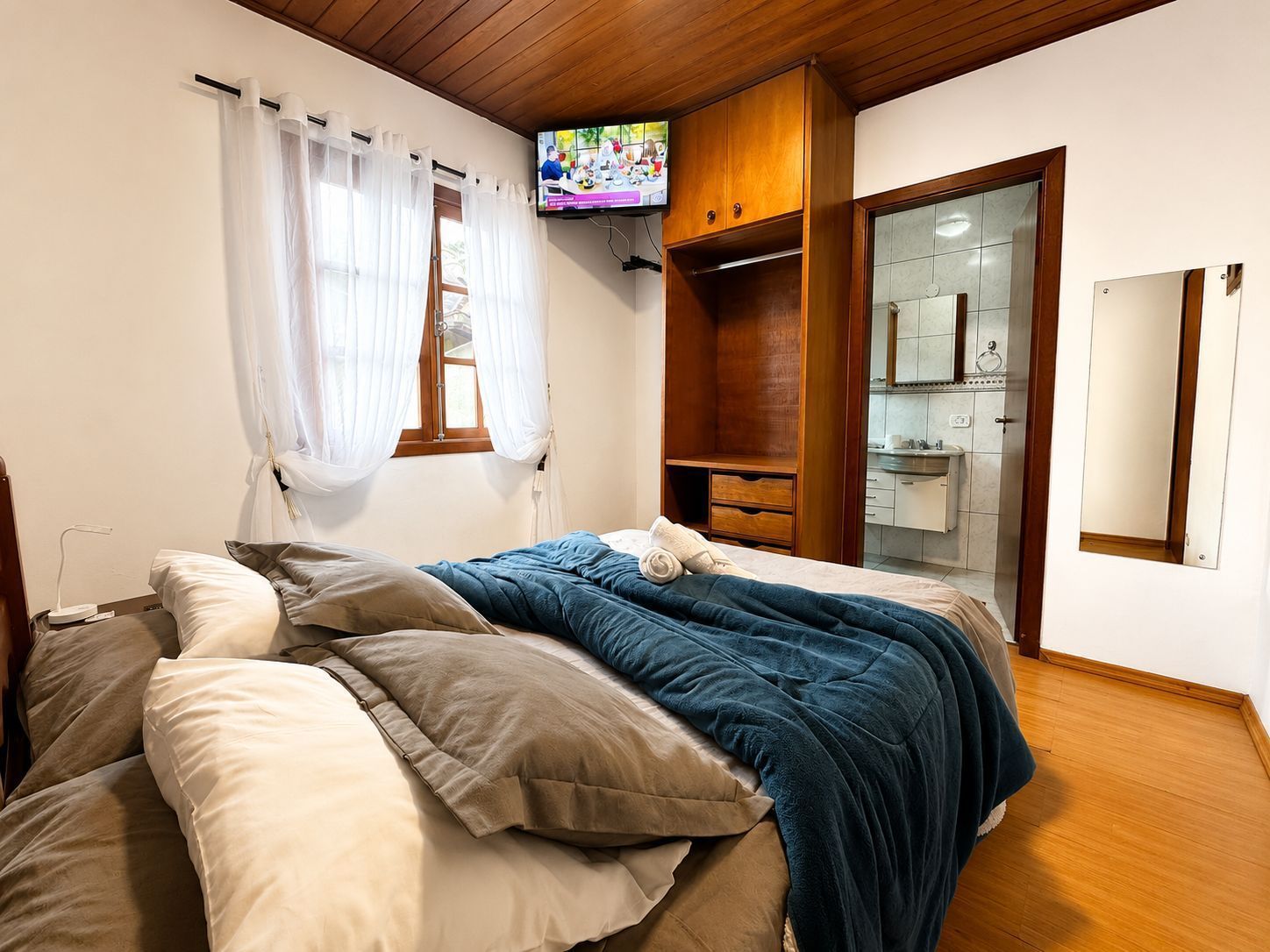 Quarto aconchegante com cama desarrumada, TV de parede e banheiro com azulejos visível através da porta aberta.