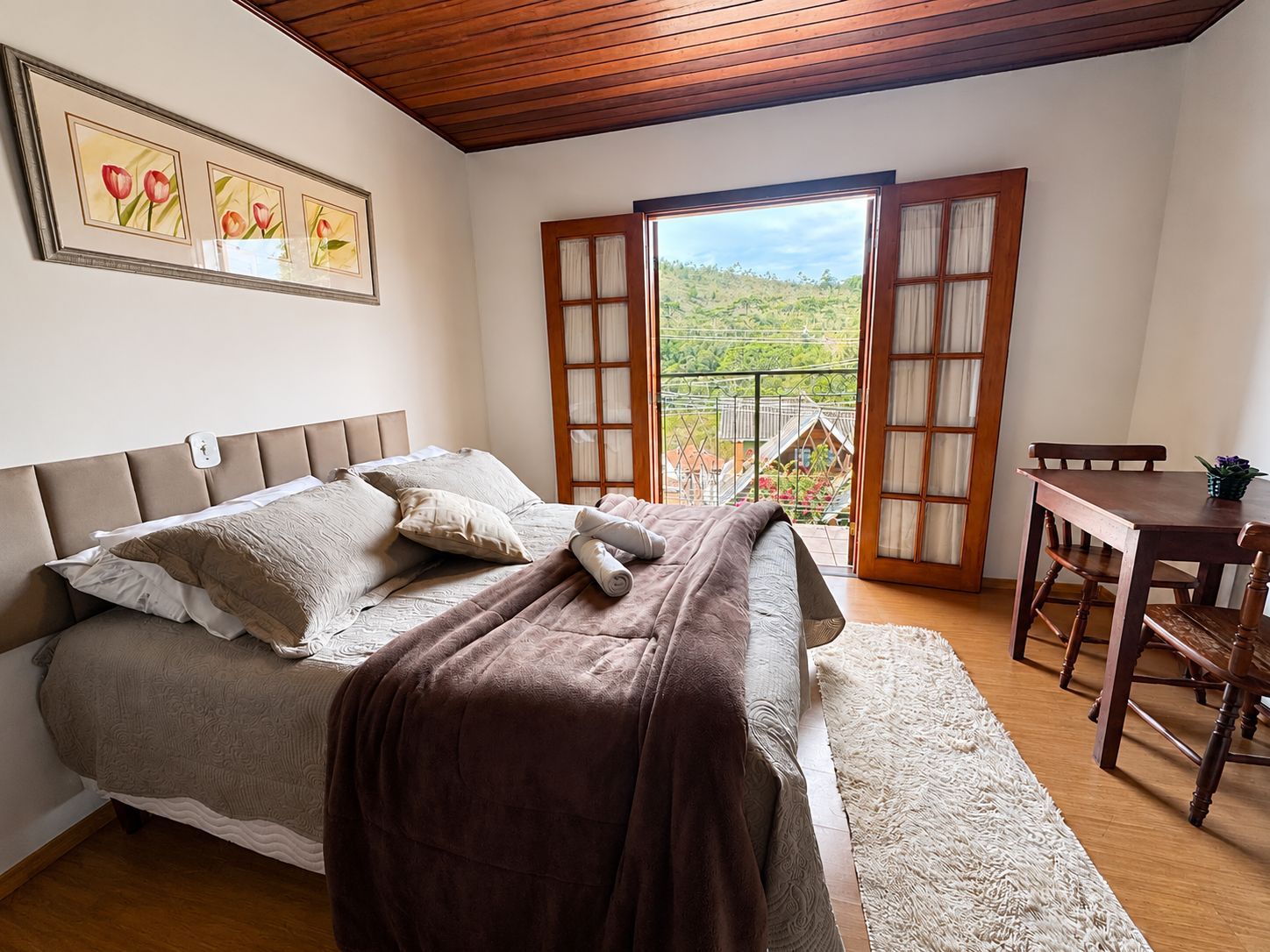 Quarto luminoso com cama, teto de madeira, escrivaninha e portas abertas para uma varanda com vista para a montanha.