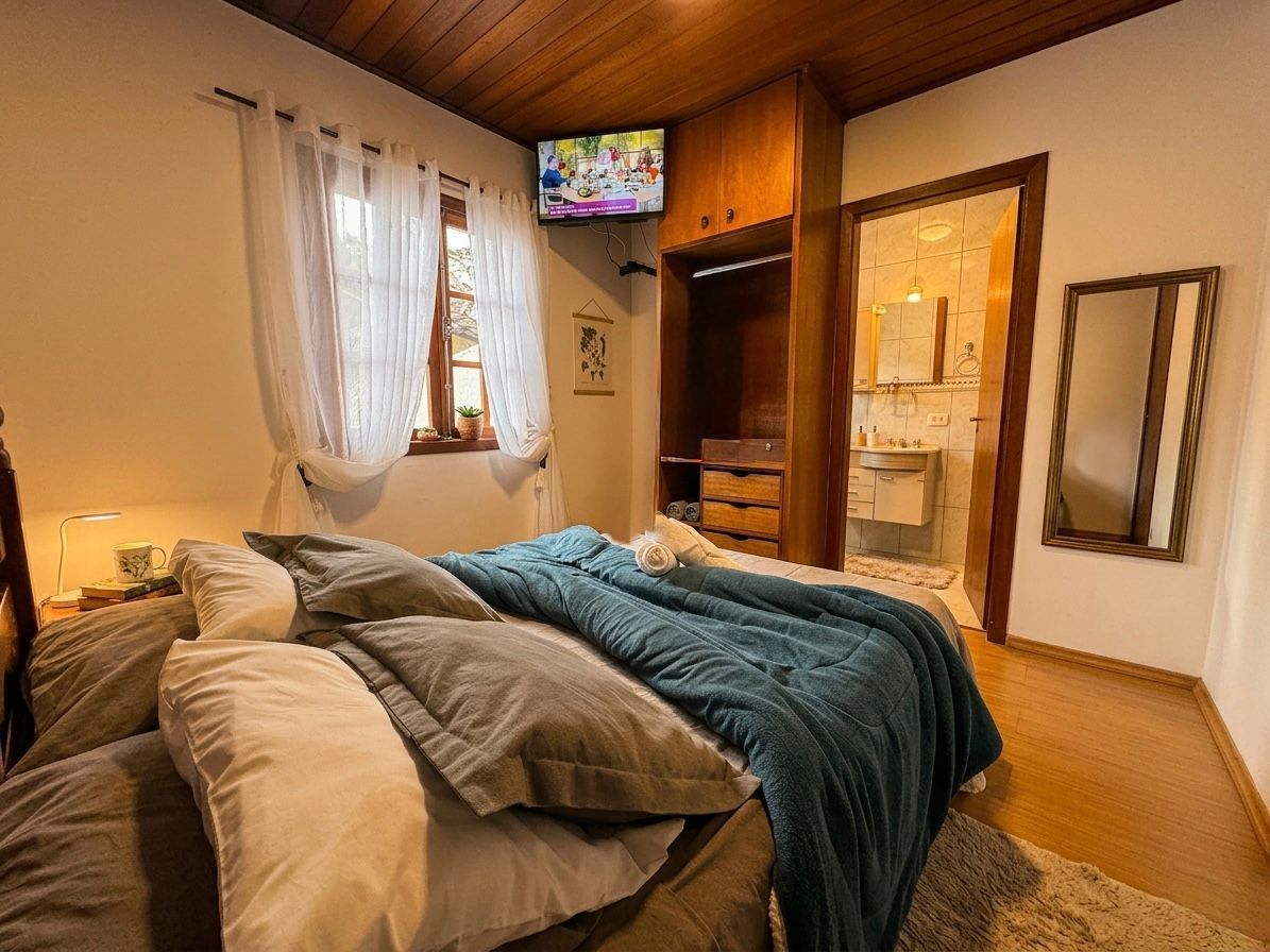 Quarto aconchegante com cama, teto de madeira, janela, espelho na parede e uma pequena TV acima da cômoda.
