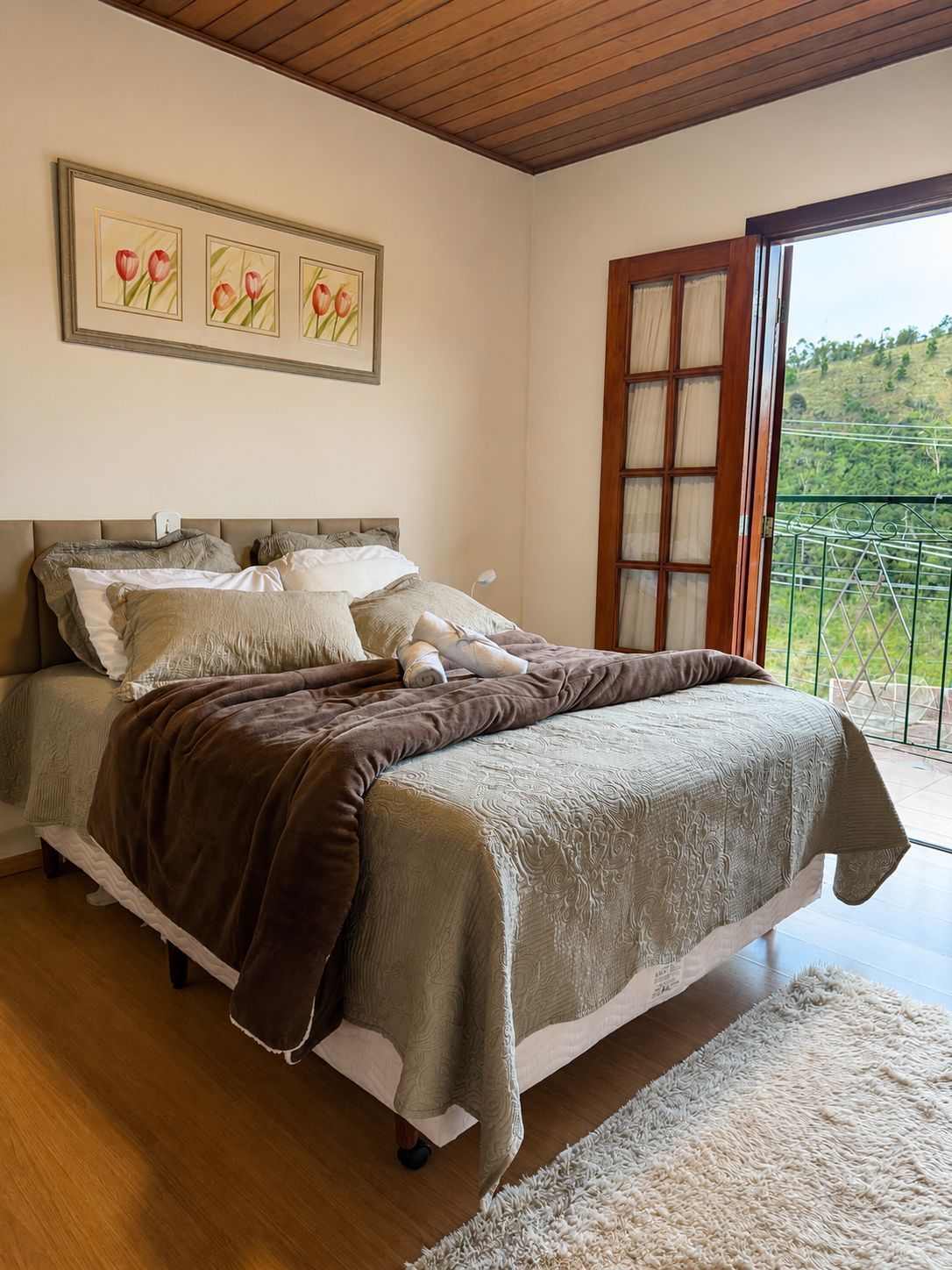 Quarto aconchegante com cama de madeira, roupa de cama em tons neutros, obras de arte emolduradas e portas abertas para uma vista verde.