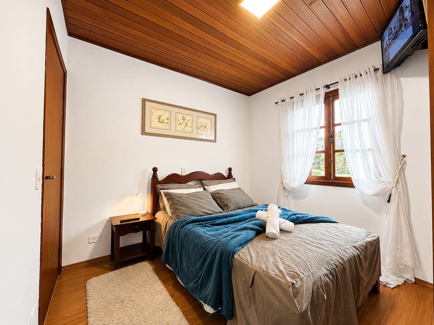 Quarto aconchegante com cama, cobertor azul-petróleo, teto de madeira e janela com cortinas translúcidas.
