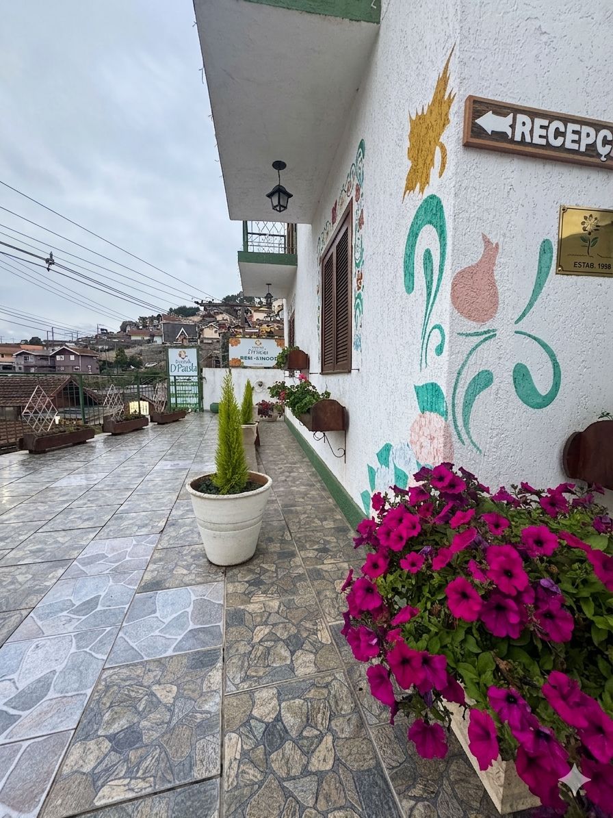 Varanda florida com piso de azulejos, plantas em vasos e vista da cidade na encosta, ao lado da placa da recepção.