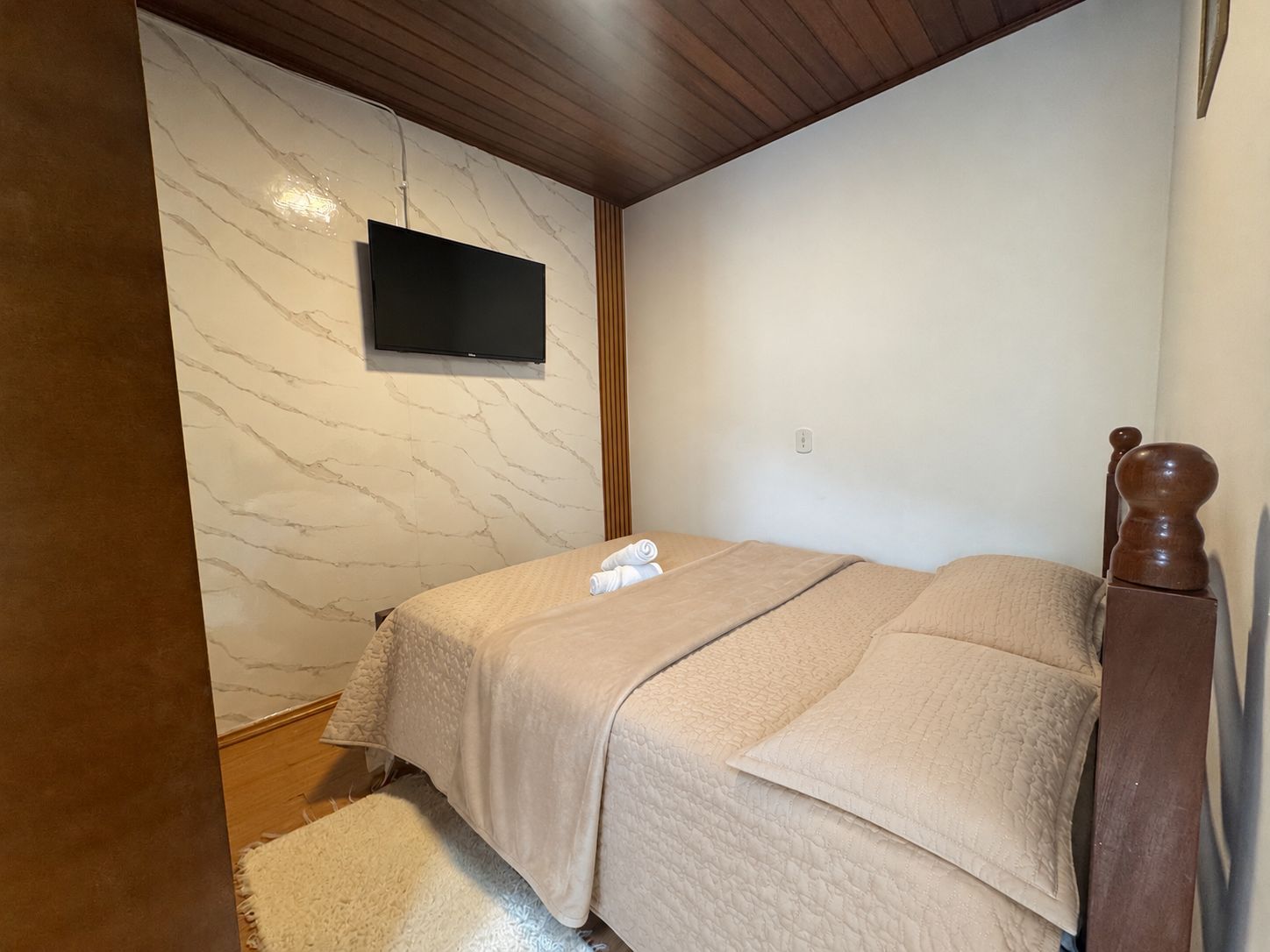 Quarto pequeno com cama arrumada, TV fixada na parede e teto com painéis de madeira.