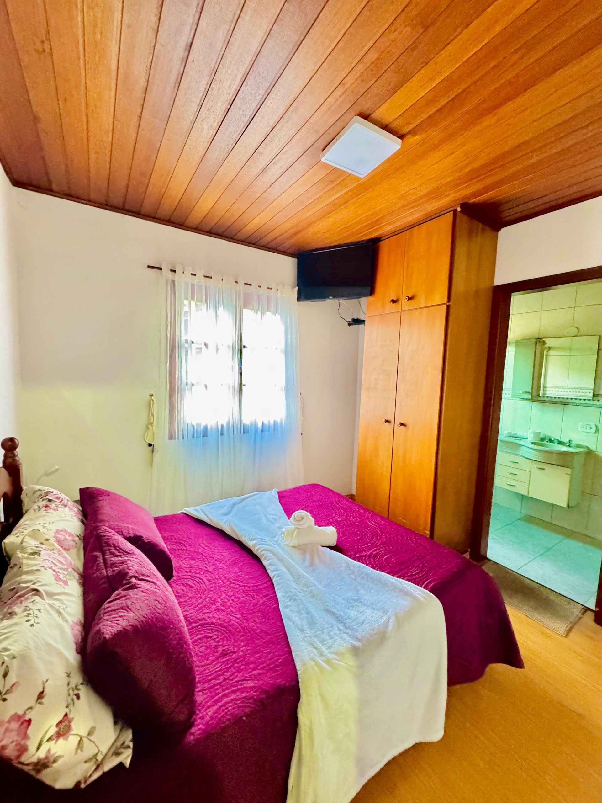 Quarto pequeno com cama, roupa de cama magenta, teto com painéis de madeira e janela com cortinas transparentes.