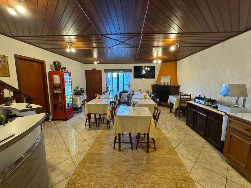 Cozinha/sala de jantar iluminada com mesas compridas, armários de madeira, piso de azulejo e uma porta azul nos fundos.