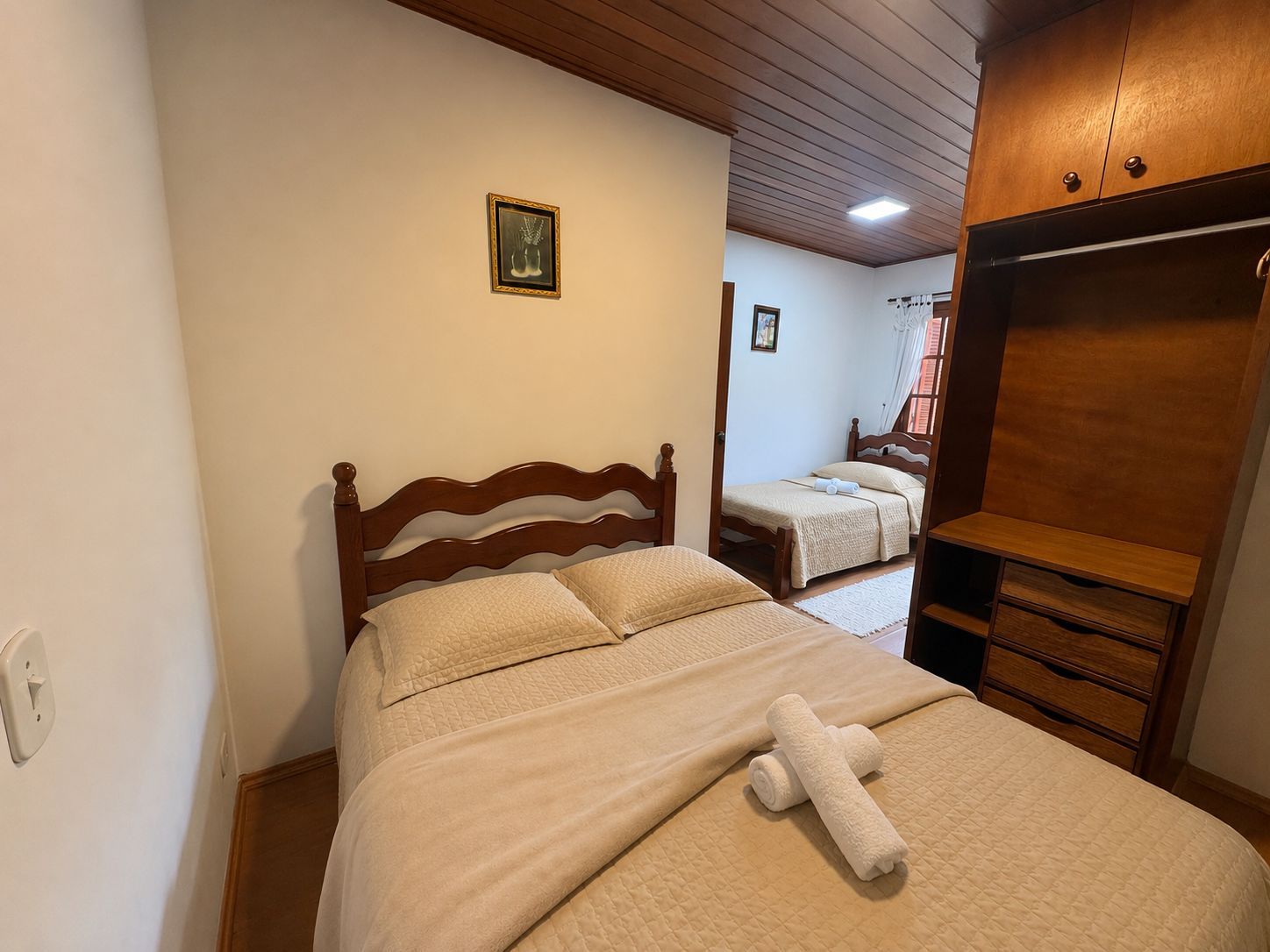 Quarto pequeno com duas camas de solteiro, móveis de madeira e uma toalha branca dobrada sobre a cama mais próxima.