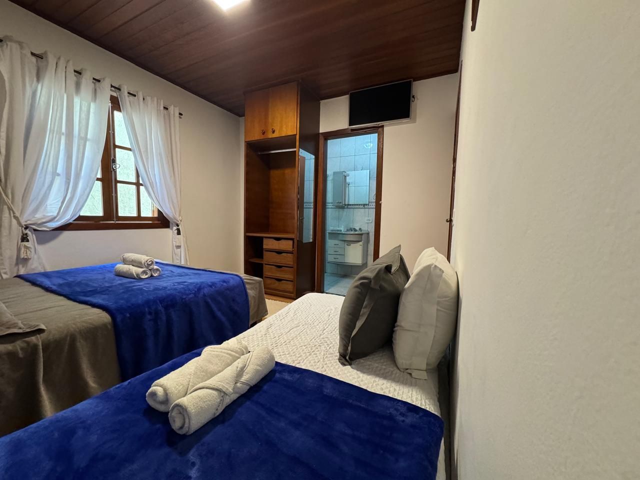 Quarto pequeno com duas camas cobertas de azul, cortinas brancas e um guarda-roupa de madeira.