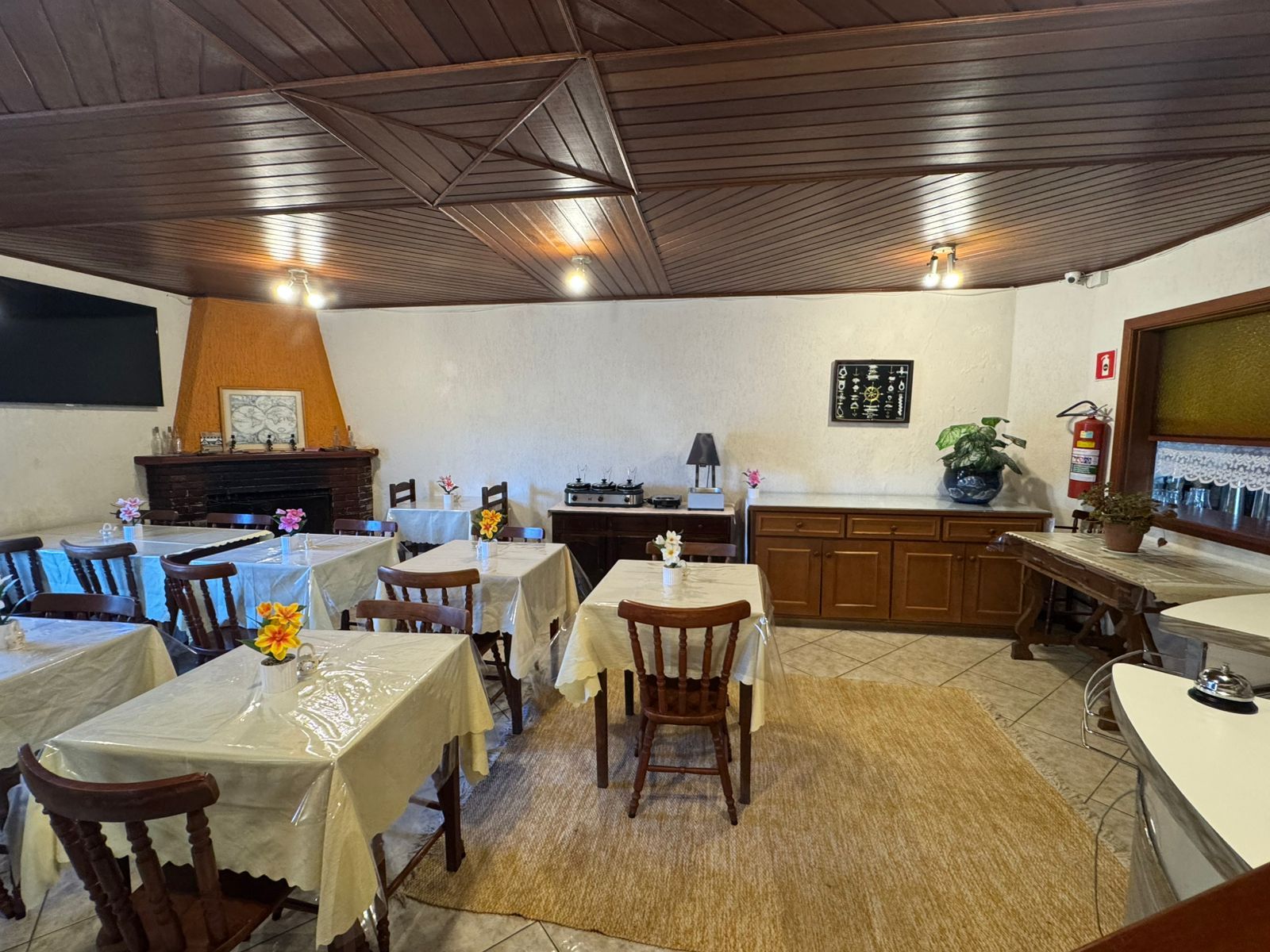 Sala de jantar com mesas cobertas com toalhas de mesa brancas, cadeiras de madeira e um longo aparador encostado na parede.