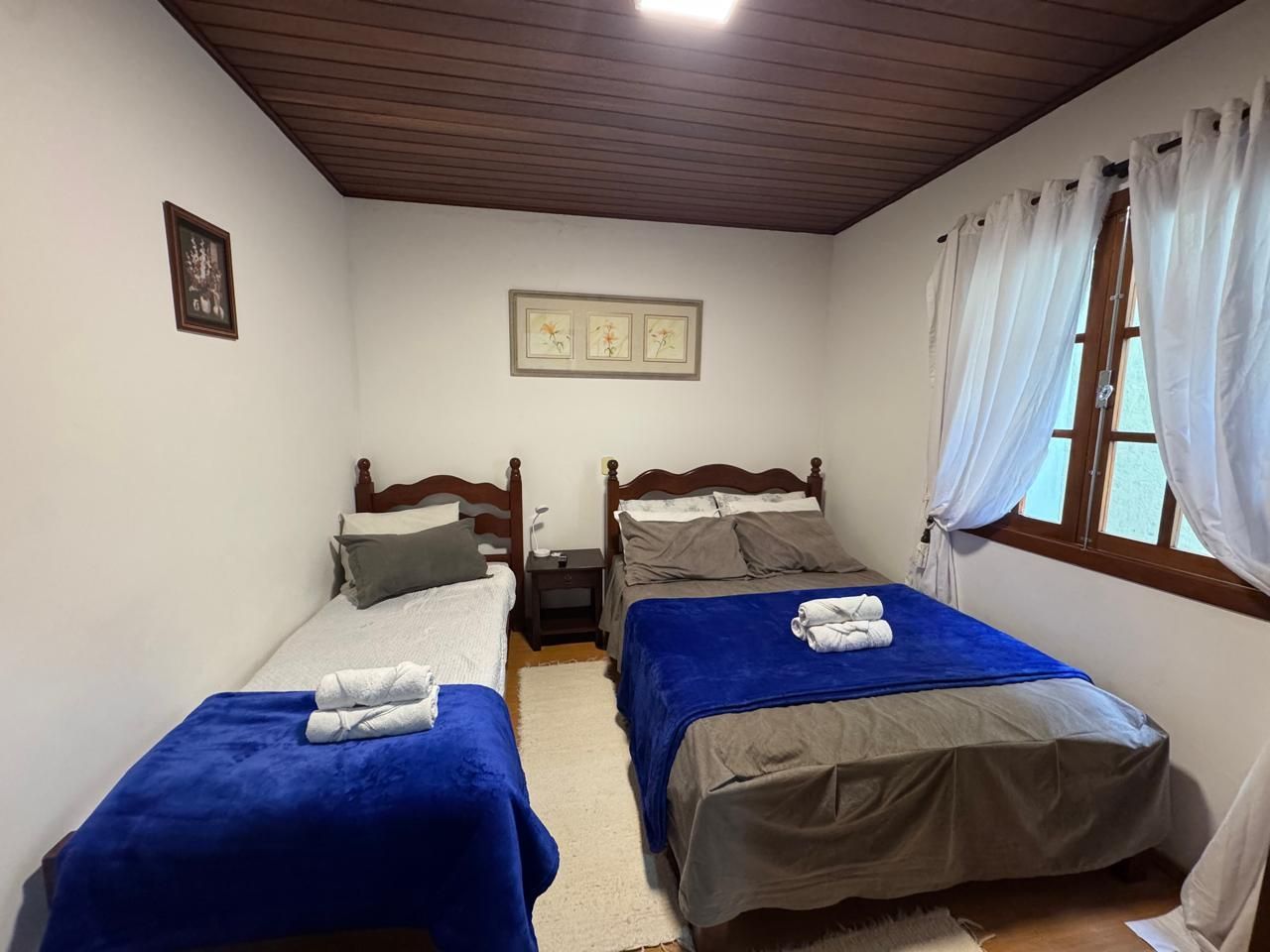 Quarto pequeno com duas camas, cobertores azuis, paredes brancas e uma janela com cortinas transparentes.