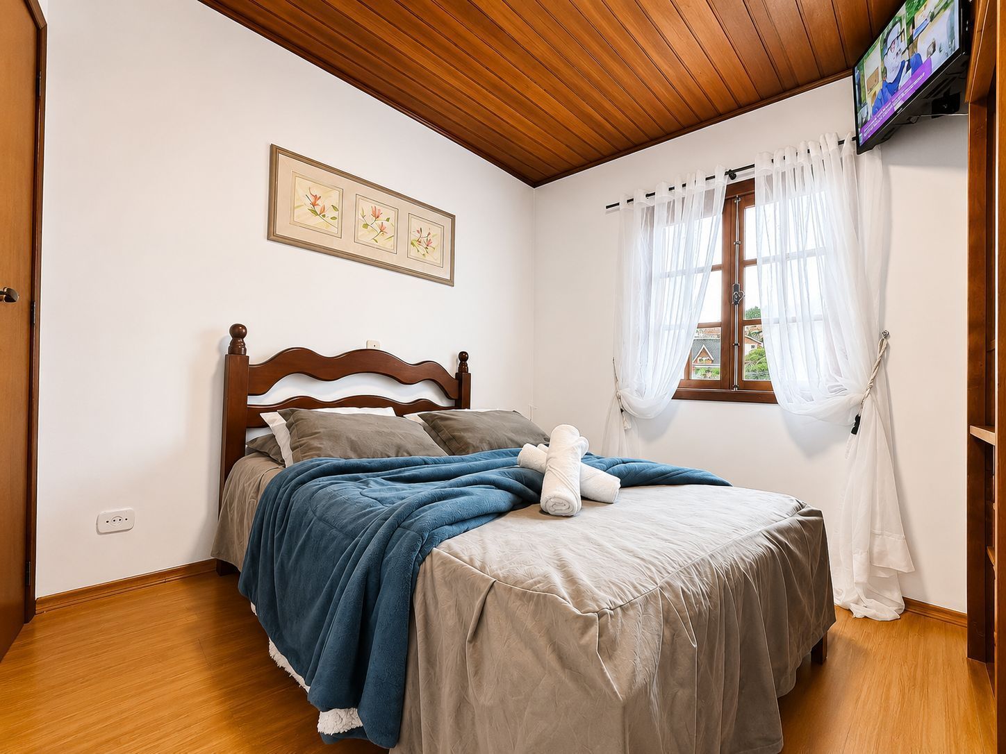 Quarto pequeno com cama de madeira, cobertor azul, edredom bege e janela com cortinas brancas.