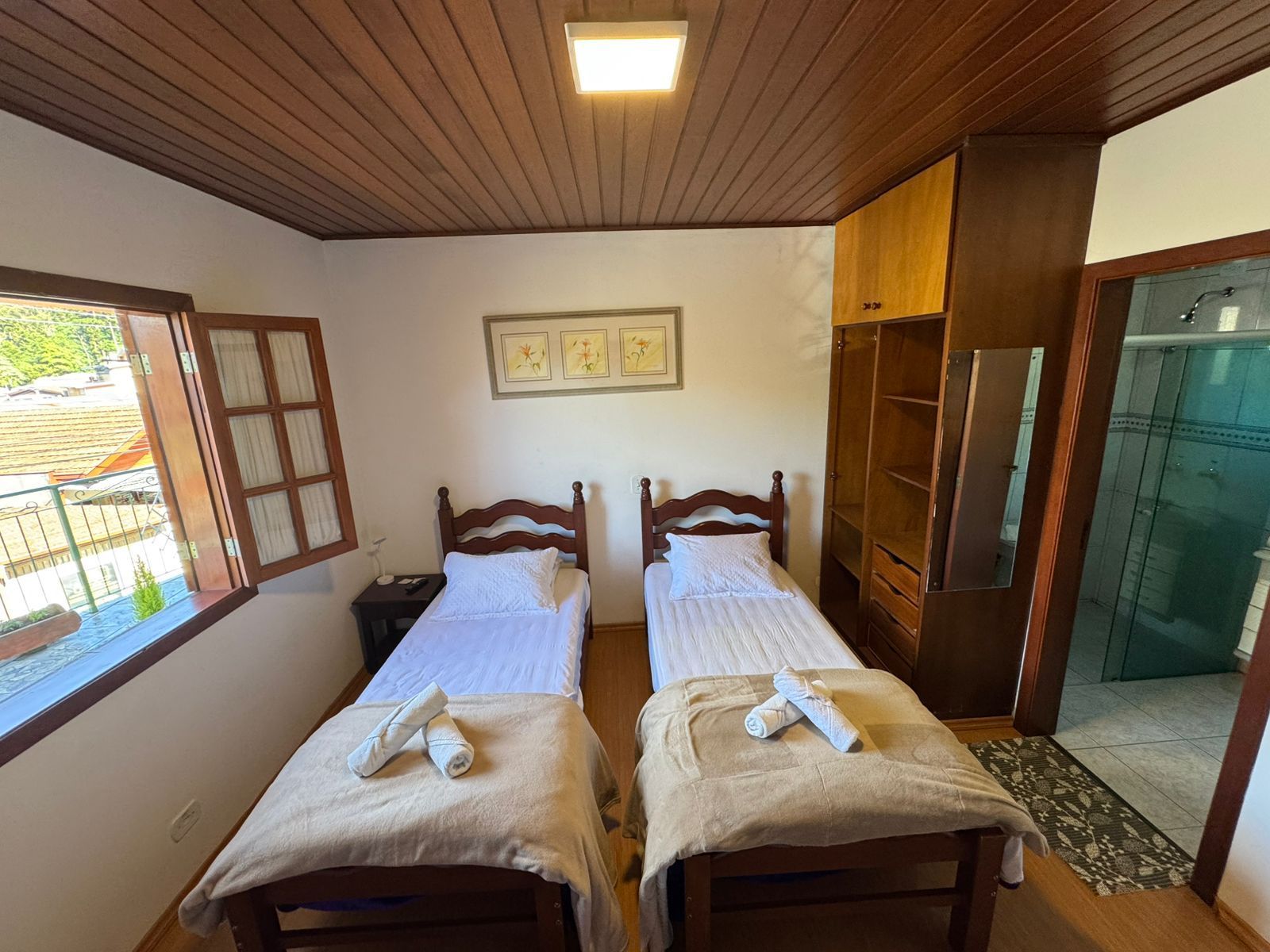 Quarto pequeno com duas camas de solteiro, teto de madeira, janela e porta para banheiro privativo.