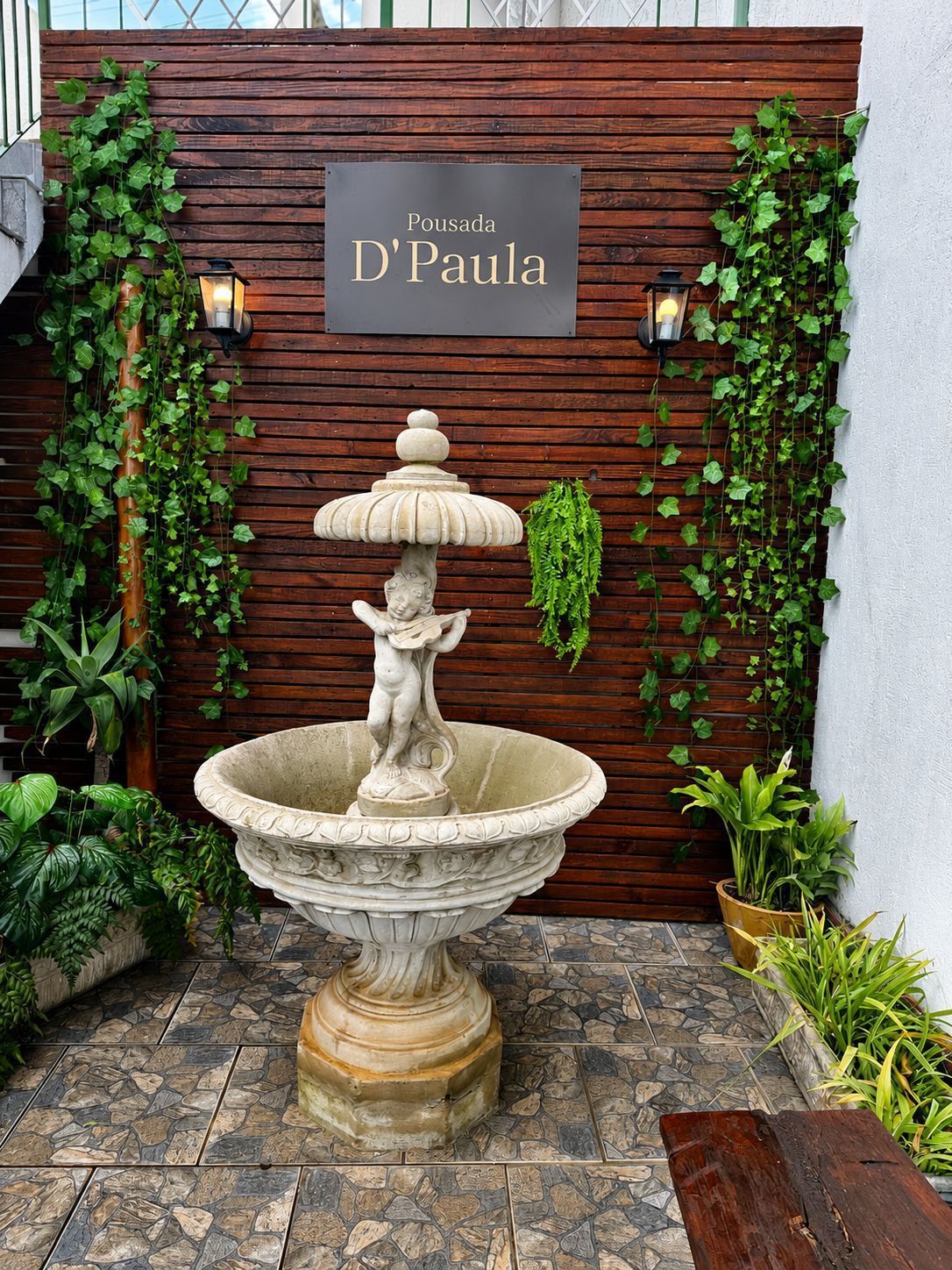 Fonte de pedra em frente a uma placa de madeira com a inscrição “D'Paula”, emoldurada por trepadeiras e plantas verdes.