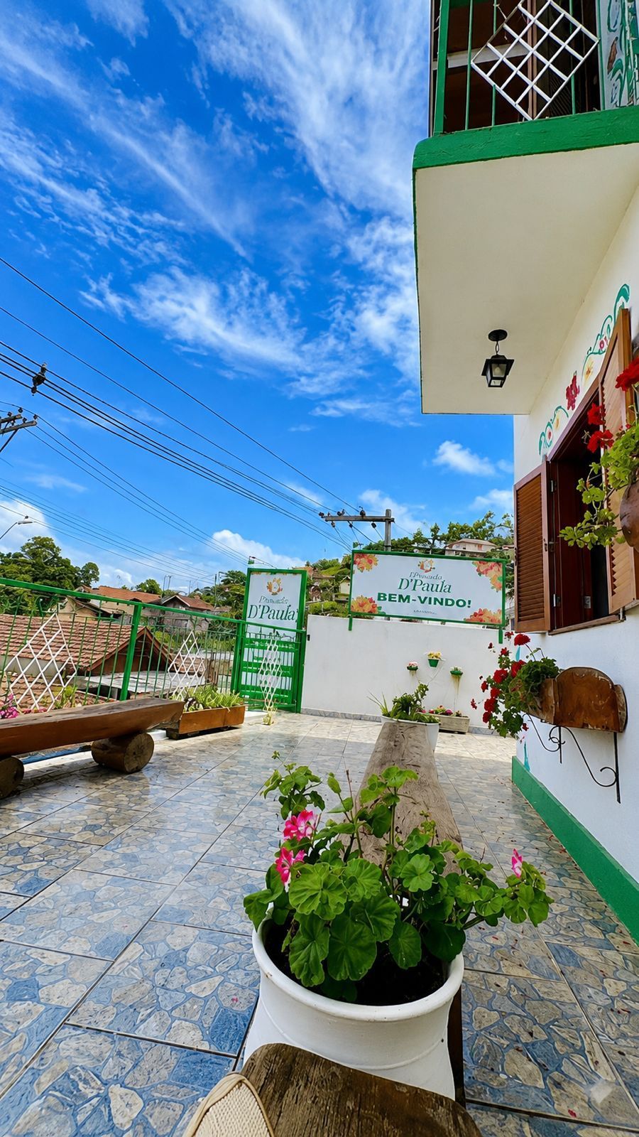 Pátio ensolarado com caminho de azulejos azuis e brancos, vasos de flores e uma placa verde ao lado de uma casa branca.