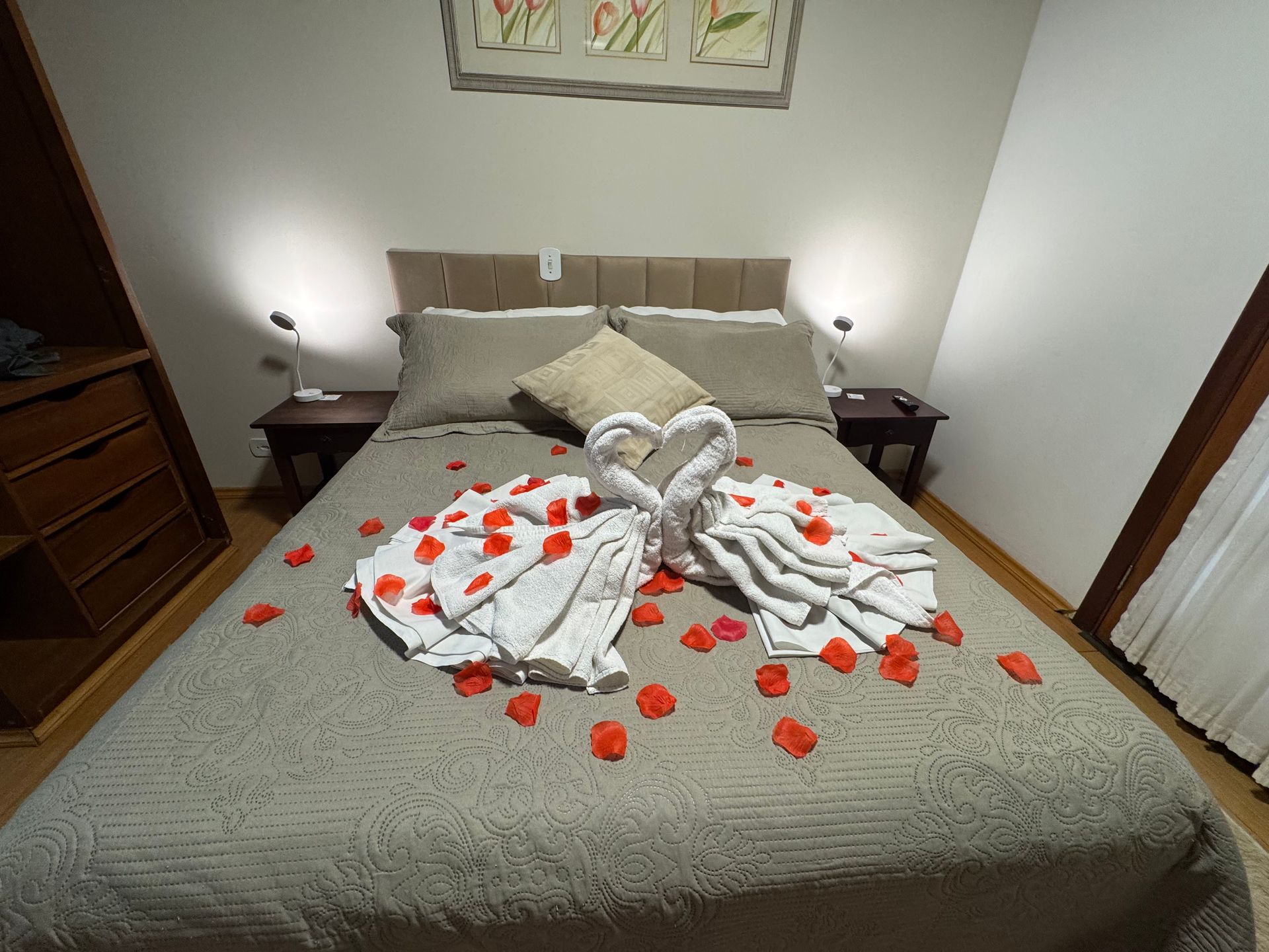 Cama de hotel decorada com cisnes feitos de toalhas e pétalas de rosas vermelhas sobre uma colcha cinza.