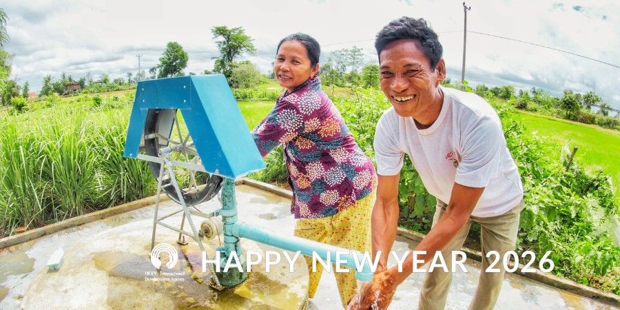 Two smiling people near a water pump in a green, outdoor setting; text reads 