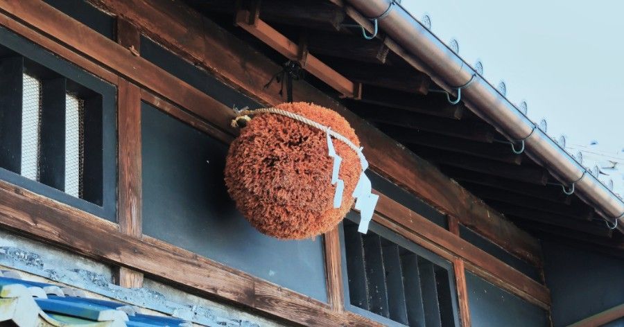 Brown spherical sugidama hanging from a wooden building, with white paper strips and a bell.
