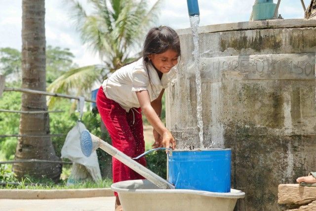 Child washing clothes with a blue bucket and basin outdoors near a concrete wall and palm tree.