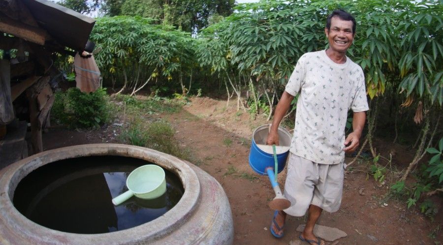 Man standing beside a circular water well, holding a blue bucket in a garden path.