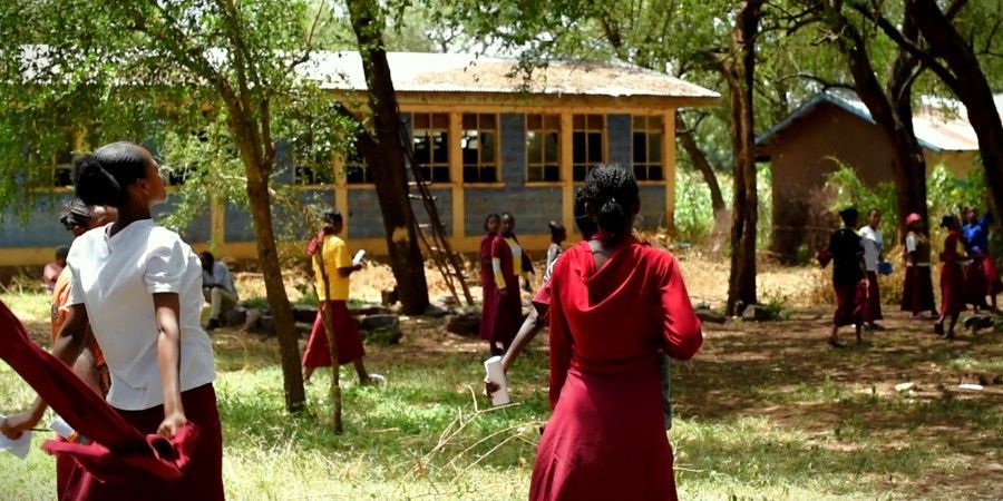 Children in red uniforms gathered outdoors near a yellow school building, some facing a tree shade.