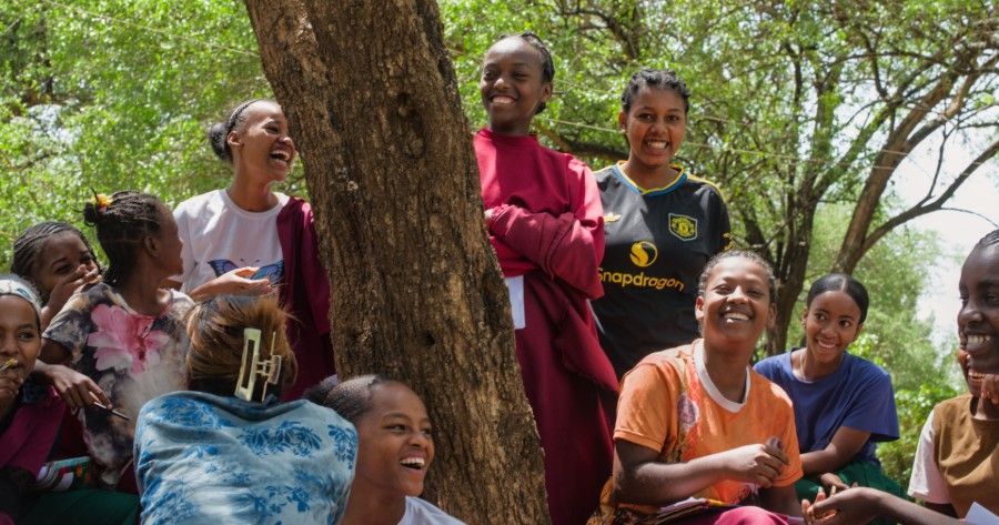 Smiling group gathered outdoors under a tree, some seated and some standing in colorful clothing.