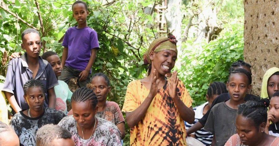 Group of people gathered outdoors among trees, with one person speaking at the center.