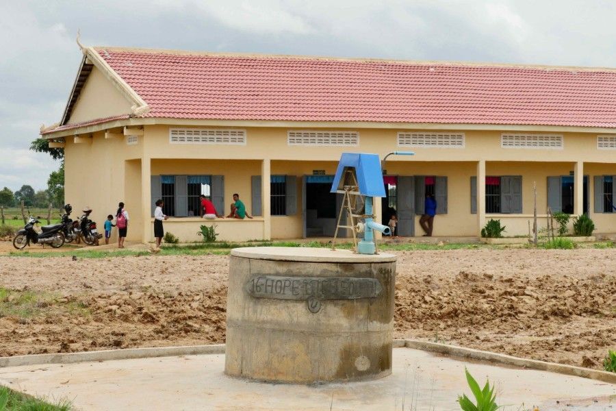 Rural school building with a water pump in the foreground and people gathered outside.