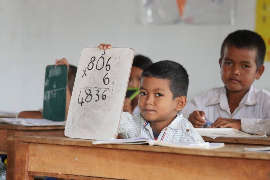 Children in a classroom, one holding a number board while seated at wooden desks