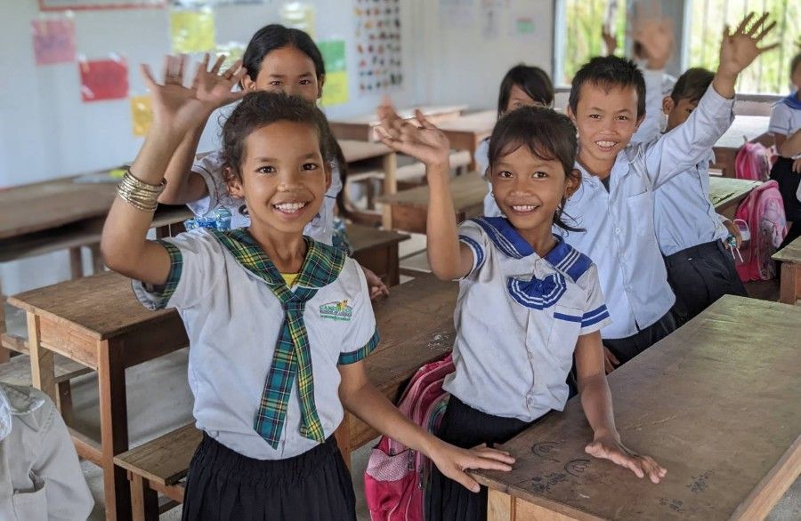 Two smiling students in uniforms wave at a classroom desk, with other children behind them