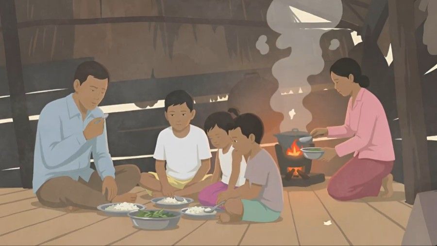 People sitting on a floor around a low meal setup near a cooking fire in a rustic wooden room
