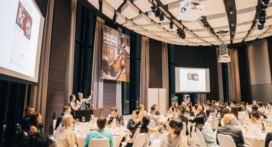 Conference audience seated in a large auditorium, watching a presentation on two projection screens.