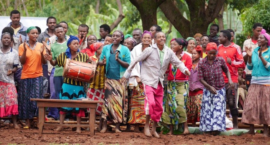 Crowd gathered outdoors around a drum, many in colorful clothing, appearing to celebrate or dance