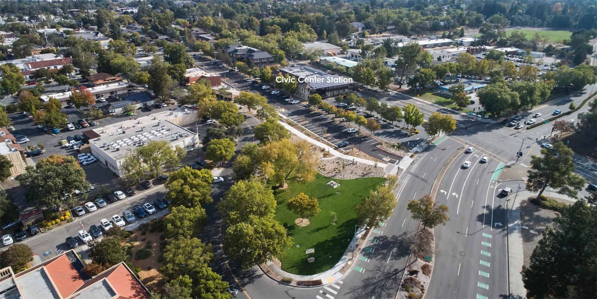 Aerial view of a city park with a green lawn, surrounded by trees, parking lots, and intersecting streets.