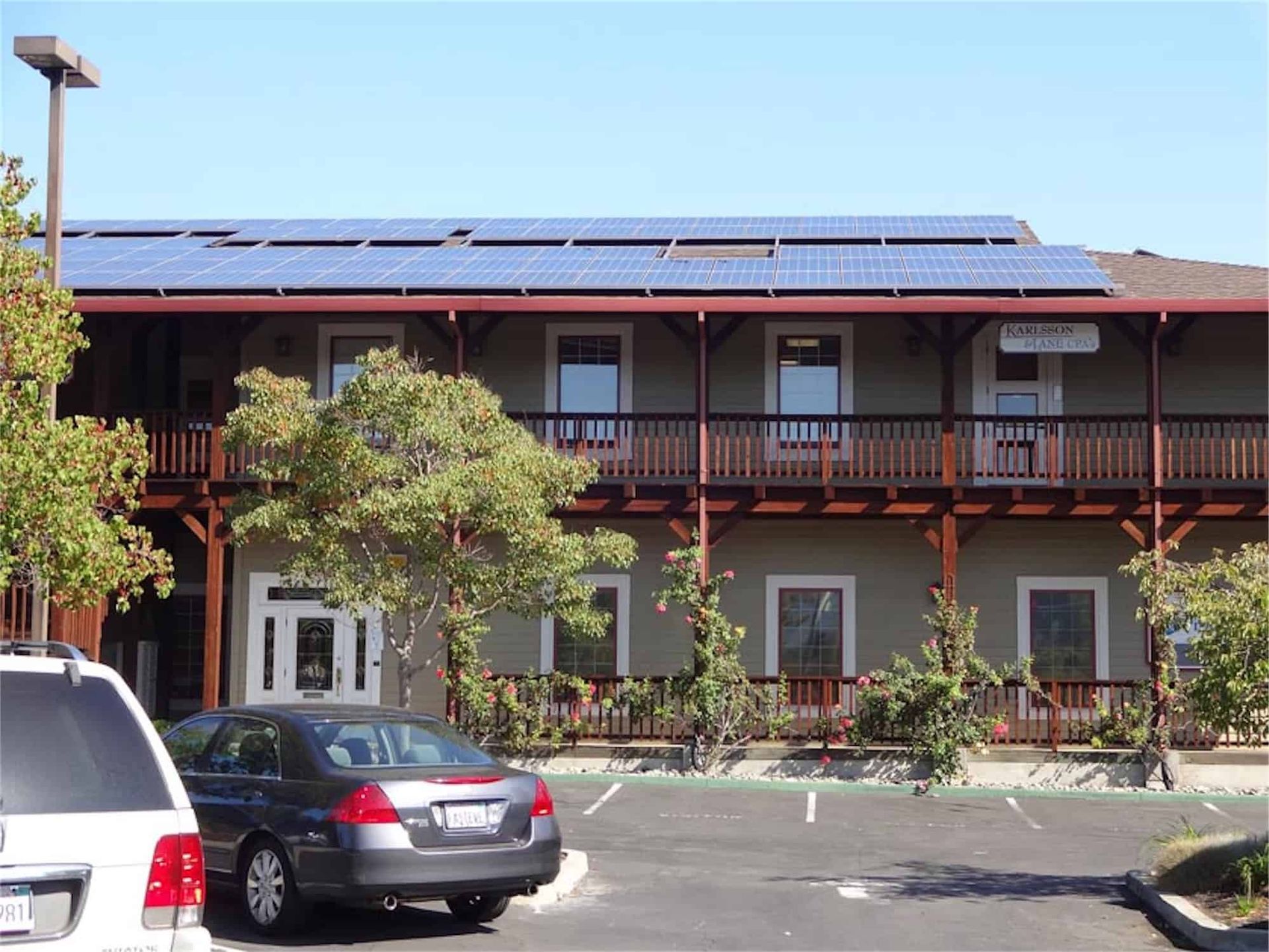 A two-story building with a wooden balcony, solar panels on the roof, and a parking lot with two cars in the foreground.
