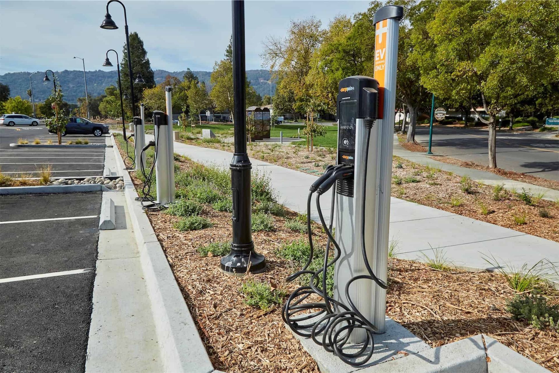 Electric vehicle charging stations in a parking lot, situated along a sidewalk with landscaping.