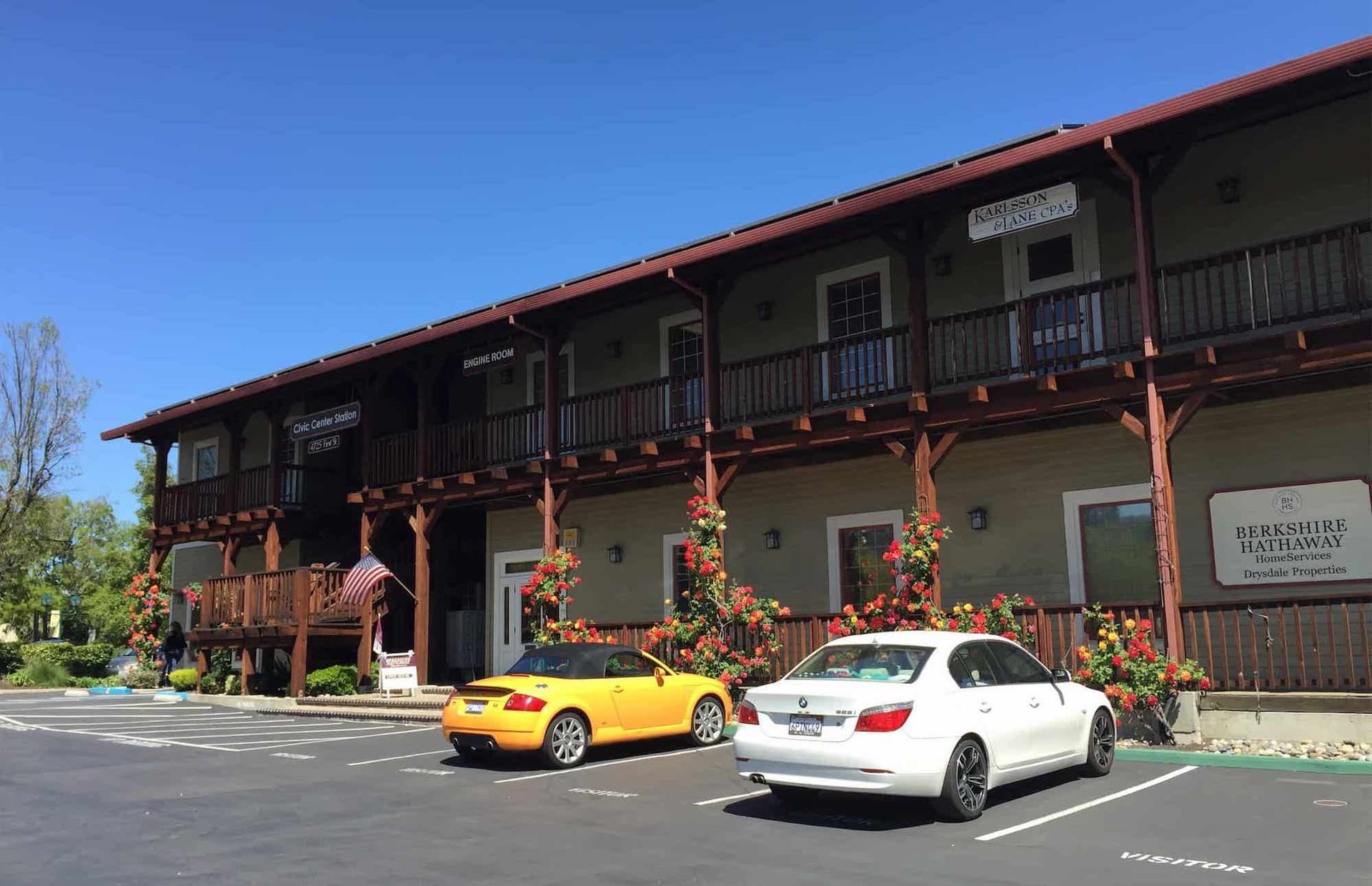 A two-story hotel with a wooden balcony and brown trim, featuring a yellow convertible and white sedan parked in front.