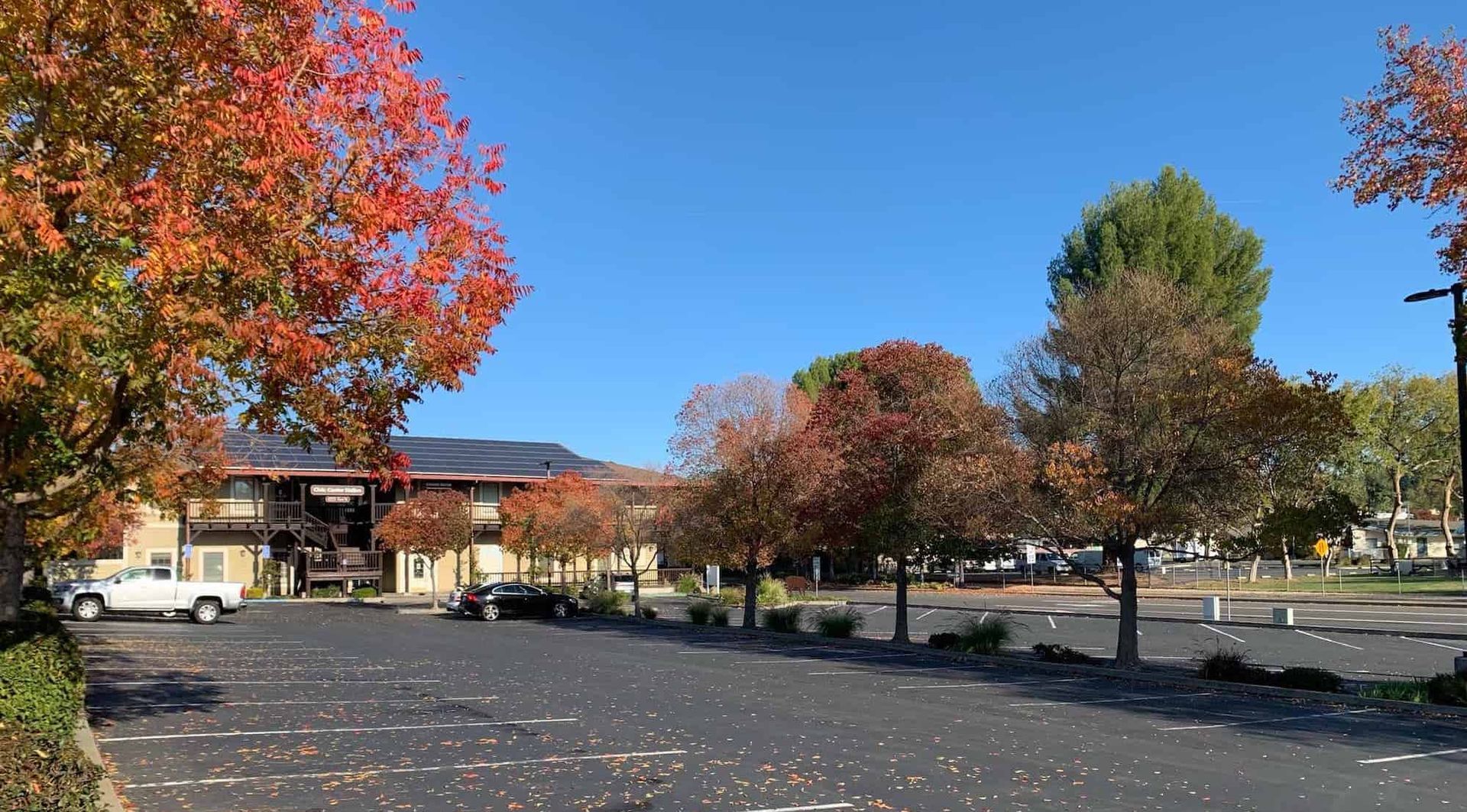 A parking lot in autumn with a two-story building in the background, surrounded by trees with orange and green leaves.