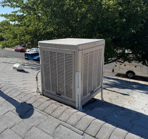 Beige rooftop HVAC unit on a shingled roof beside trees and parked cars.