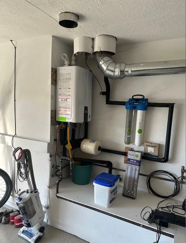 Water heater and furnace setup in a garage, with ducts, pipes, and utility equipment along the wall.